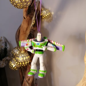 Peut inclure: Ornement de No&euml;l Buzz l'&Eacute;clair, avec une combinaison spatiale blanche, verte et violette. L'ornement est suspendu par un ruban violet. La figurine a les bras tendus et pointe. Des boules d&eacute;coratives dor&eacute;es sont en arri&egrave;re-plan.