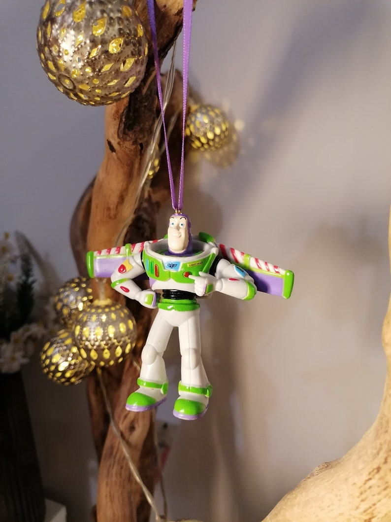 Peut inclure: Ornement de No&euml;l Buzz l'&Eacute;clair, avec une combinaison spatiale blanche, verte et violette. L'ornement est suspendu par un ruban violet. La figurine a les bras tendus et pointe. Des boules d&eacute;coratives dor&eacute;es sont en arri&egrave;re-plan.