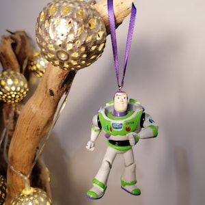 May include: A green and white plastic toy figurine of Buzz Lightyear from the Toy Story movie franchise hanging from a purple ribbon. The figurine is wearing a green spacesuit with a white chest plate and a purple and white emblem on the chest. The figurine is holding a laser gun in its right hand.