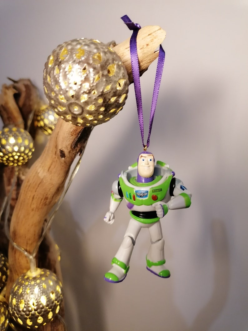 May include: A green and white plastic toy figurine of Buzz Lightyear from the Toy Story movie franchise hanging from a purple ribbon. The figurine is wearing a green spacesuit with a white chest plate and a purple and white emblem on the chest. The figurine is holding a laser gun in its right hand.