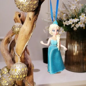 K&ouml;nnte beinhalten: Eine blaue und wei&szlig;e Plastikfigur einer Prinzessin mit blonden Haaren, die an einem blauen Band an einem Holzast mit silbernen und goldenen Ornamenten h&auml;ngt.