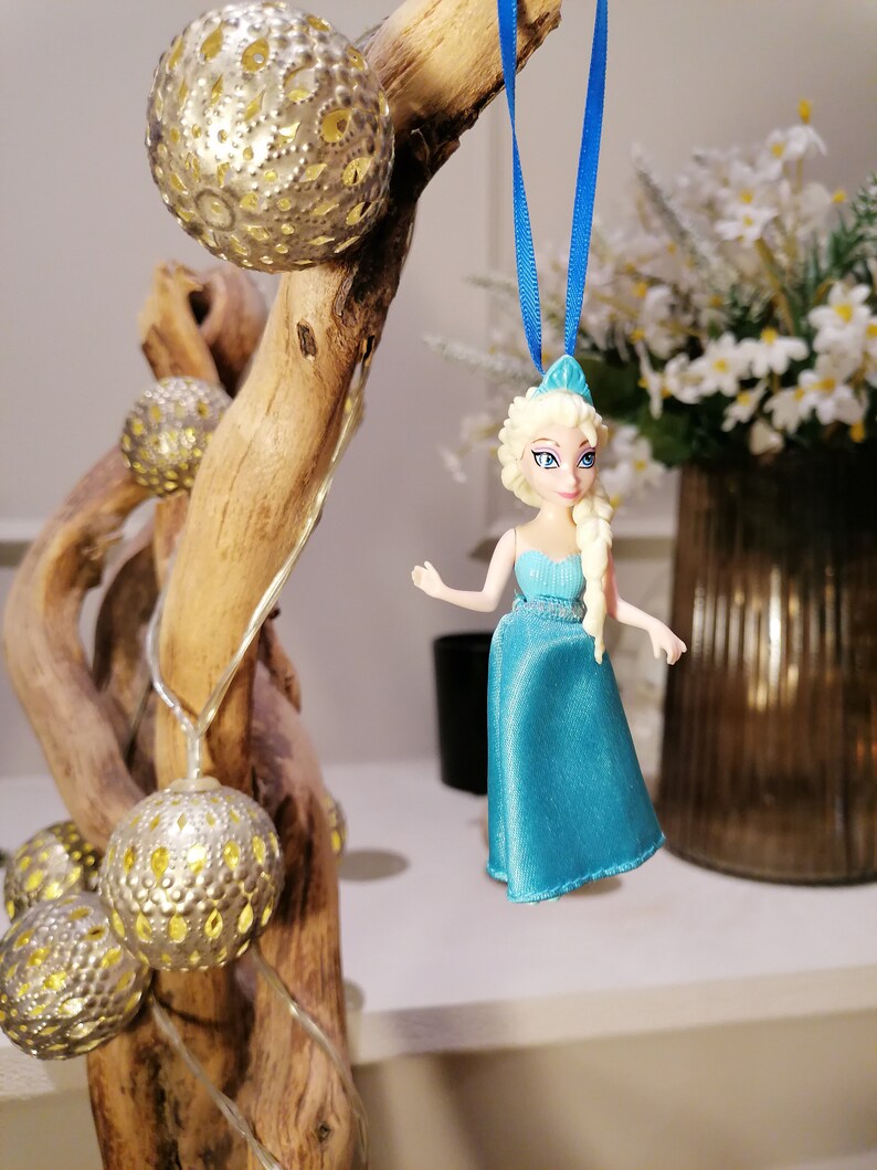 K&ouml;nnte beinhalten: Eine blaue und wei&szlig;e Plastikfigur einer Prinzessin mit blonden Haaren, die an einem blauen Band an einem Holzast mit silbernen und goldenen Ornamenten h&auml;ngt.