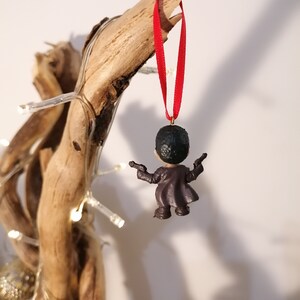 May include: A small, dark-haired figurine wearing a brown coat and holding two guns. The figurine is hanging from a red ribbon on a piece of driftwood.