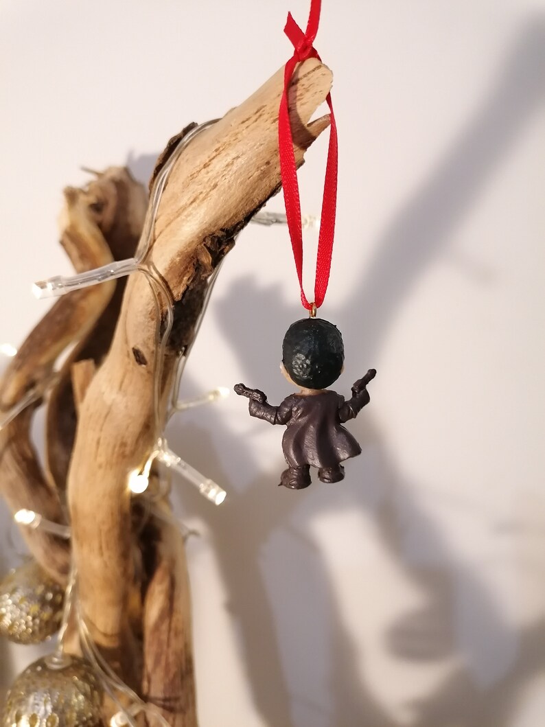 May include: A small, dark-haired figurine wearing a brown coat and holding two guns. The figurine is hanging from a red ribbon on a piece of driftwood.
