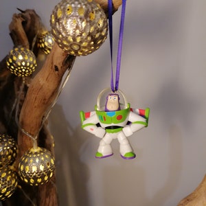 Puede incluir: Un adorno de plástico morado y blanco de Buzz Lightyear de la franquicia Toy Story. El adorno está colgado de una cinta morada en una rama con luces decorativas doradas y blancas.