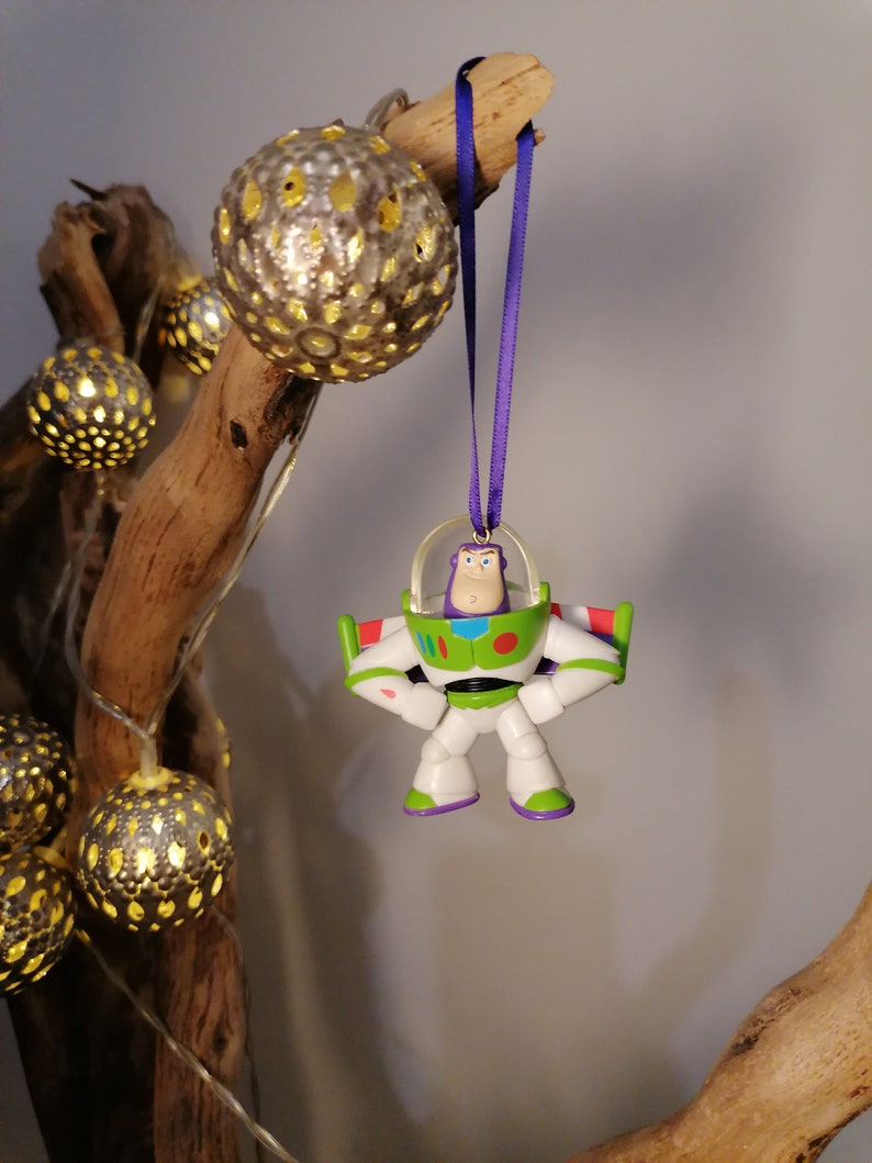 Puede incluir: Un adorno de pl&aacute;stico morado y blanco de Buzz Lightyear de la franquicia Toy Story. El adorno est&aacute; colgado de una cinta morada en una rama con luces decorativas doradas y blancas.