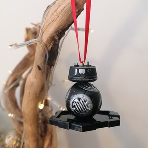 K&ouml;nnte beinhalten: Schwarzer und silberner Star Wars BB-8 Weihnachtsbaumschmuck mit einem roten Band, das an einem Ast h&auml;ngt.