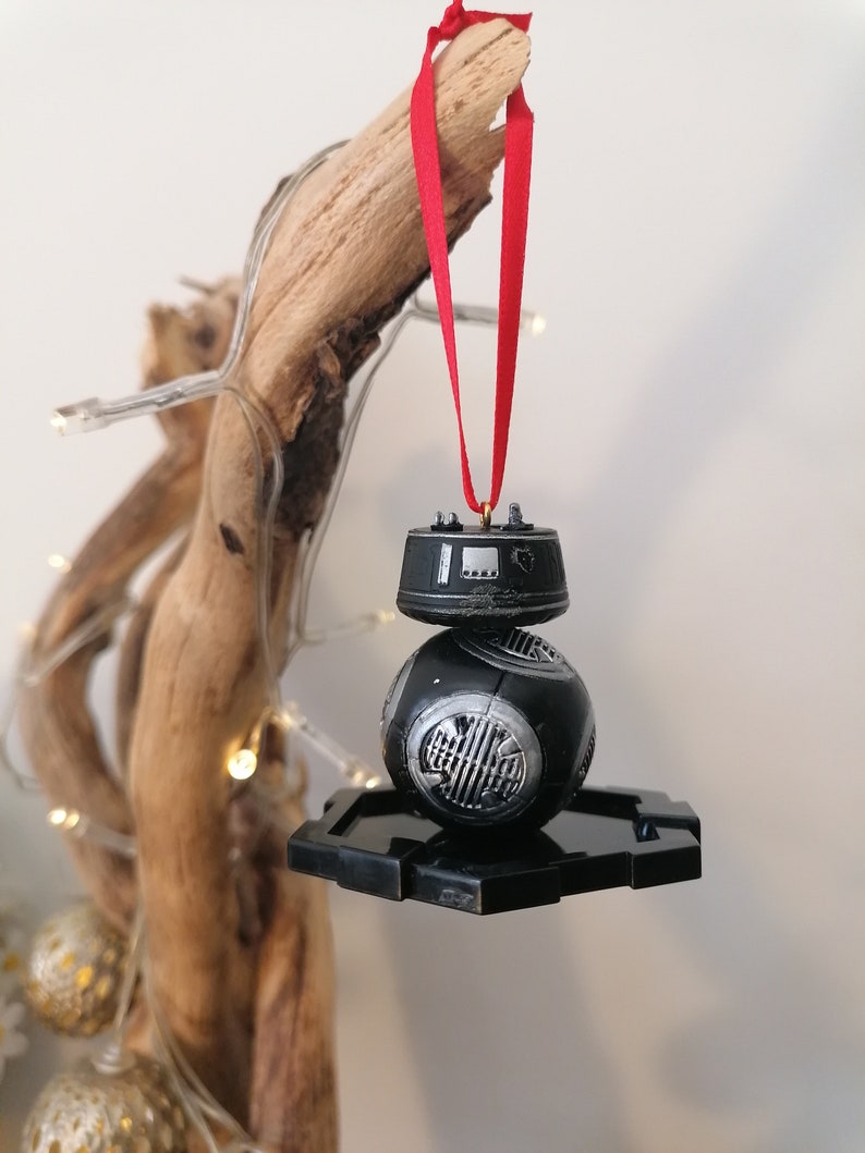 K&ouml;nnte beinhalten: Schwarzer und silberner Star Wars BB-8 Weihnachtsbaumschmuck mit einem roten Band, das an einem Ast h&auml;ngt.