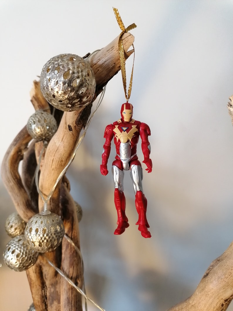 Pode incluir: Um ornamento de figura de a&ccedil;&atilde;o do Homem de Ferro vermelho e prateado pendurado em um galho de madeira flutuante com um cord&atilde;o dourado. O ornamento &eacute; decorado com um traje vermelho e prateado e um capacete vermelho com uma m&aacute;scara facial amarela.