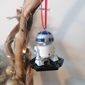 May include: A white and blue R2-D2 Star Wars character ornament hanging from a red ribbon on a wooden branch. The ornament is attached to a black base.