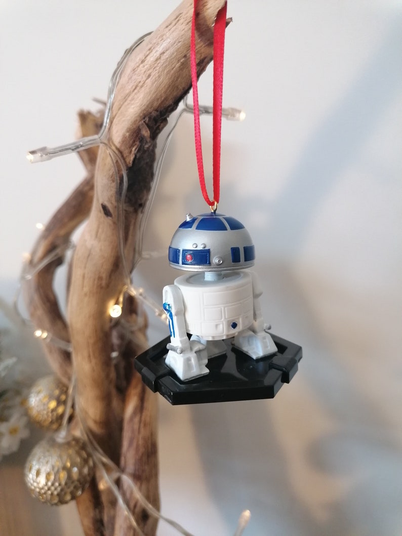 May include: A white and blue R2-D2 Star Wars character ornament hanging from a red ribbon on a wooden branch. The ornament is attached to a black base.