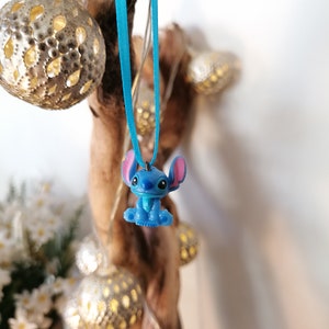 May include: A blue Stitch character charm hanging from a turquoise ribbon. The charm is a 3D representation of the character from the Disney movie Lilo & Stitch.