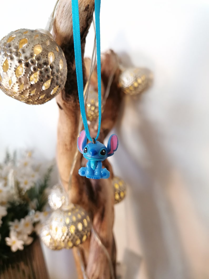 May include: A blue Stitch character charm hanging from a turquoise ribbon. The charm is a 3D representation of the character from the Disney movie Lilo & Stitch.