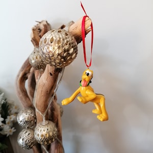 Op de afbeelding: Een gele cartoon-hond kerstversiering hangend aan een rood lint aan een tak met gouden en zilveren ornamenten.