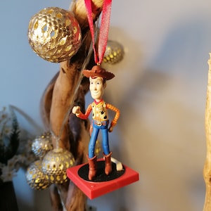 Puede incluir: Una figura de vaquero roja y blanca con un sombrero marr&oacute;n y pantalones vaqueros azules, de pie sobre una plataforma roja. La figura est&aacute; colgada de una cinta roja.
