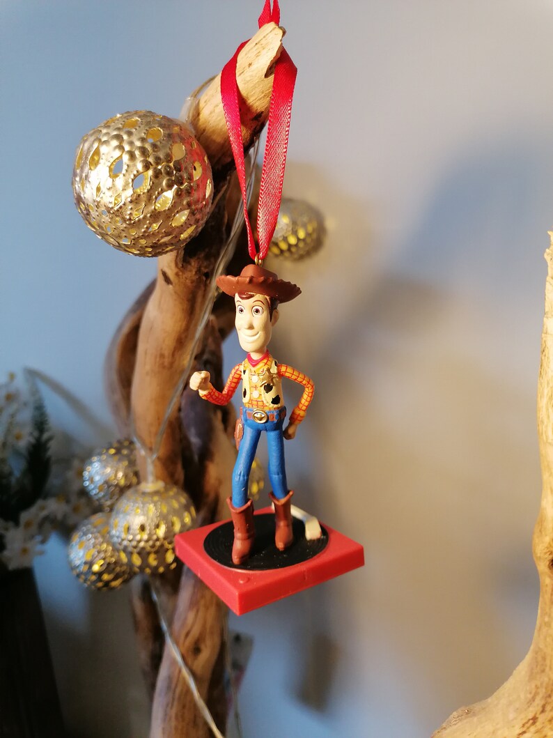Puede incluir: Una figura de vaquero roja y blanca con un sombrero marr&oacute;n y pantalones vaqueros azules, de pie sobre una plataforma roja. La figura est&aacute; colgada de una cinta roja.