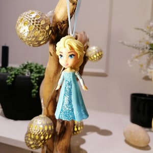 May include: A blue and white figurine of a girl with blonde hair wearing a blue dress with glitter. The figurine is hanging from a branch with a blue ribbon.