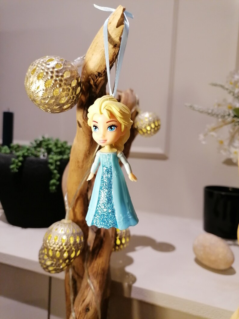 May include: A blue and white figurine of a girl with blonde hair wearing a blue dress with glitter. The figurine is hanging from a branch with a blue ribbon.