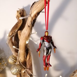May include: A red and gold Iron Man Christmas ornament hanging from a branch with red ribbon. The ornament is a detailed figurine of Iron Man in flight.