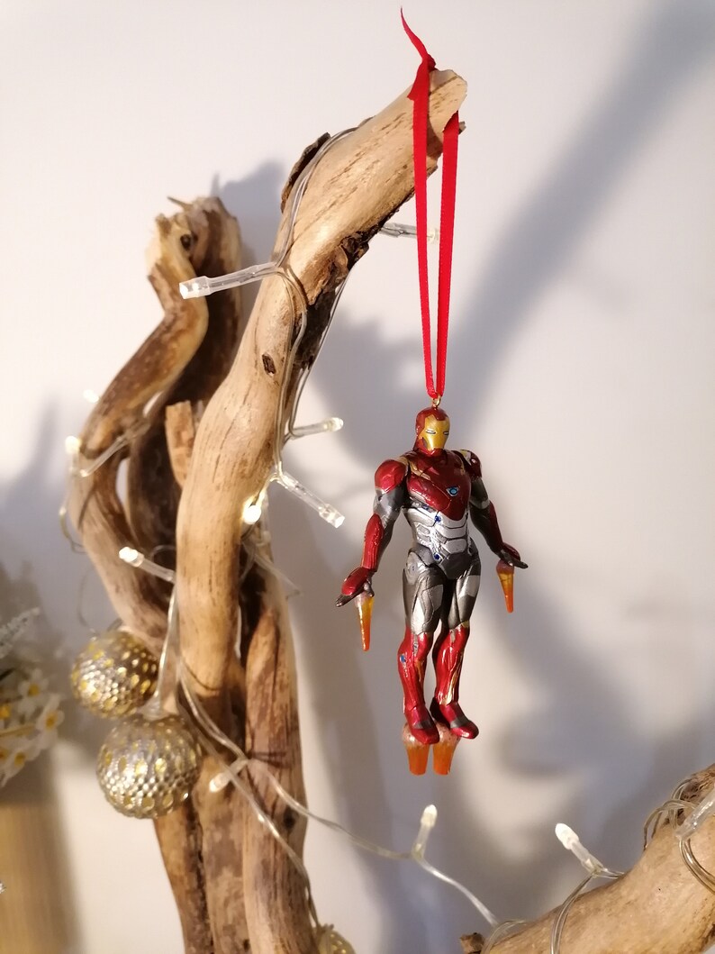 May include: A red and gold Iron Man Christmas ornament hanging from a branch with red ribbon. The ornament is a detailed figurine of Iron Man in flight.