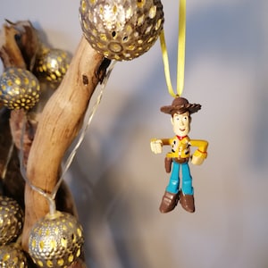 May include: A decorative Christmas ornament featuring a Woody action figure from Toy Story, suspended by a yellow ribbon. The ornament is set against a backdrop of a wooden branch adorned with string lights and silver, spherical ornaments.