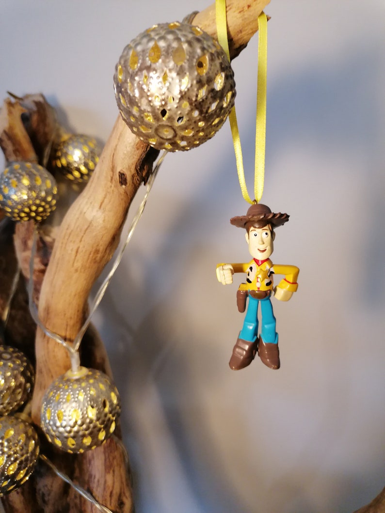 May include: A decorative Christmas ornament featuring a Woody action figure from Toy Story, suspended by a yellow ribbon. The ornament is set against a backdrop of a wooden branch adorned with string lights and silver, spherical ornaments.