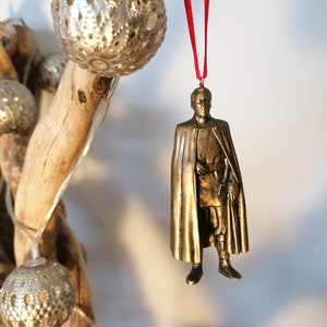 May include: A gold-colored ornament depicting a man in a long coat and hat. The ornament is hanging from a red ribbon.