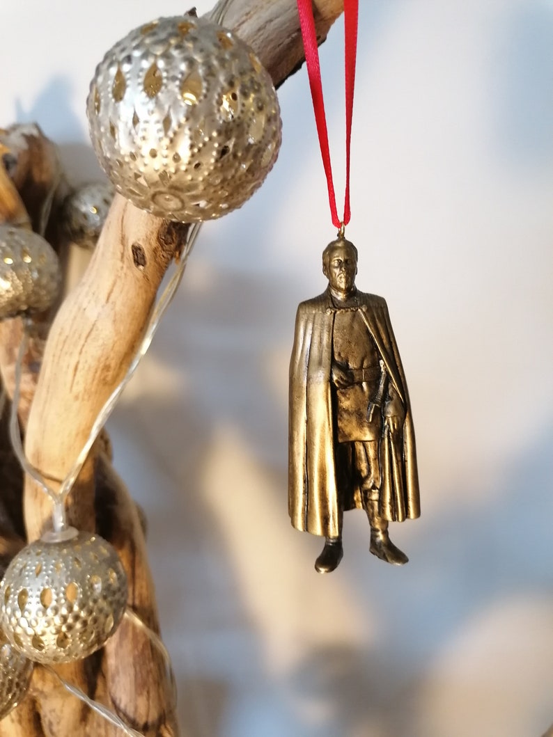 May include: A gold-colored ornament depicting a man in a long coat and hat. The ornament is hanging from a red ribbon.
