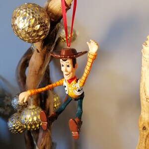 May include: A red-stringed ornament of Woody from Toy Story, wearing his signature cowboy hat and vest, with a yellow shirt and blue jeans. He is posed with his arms outstretched and legs bent, as if he is jumping or flying.