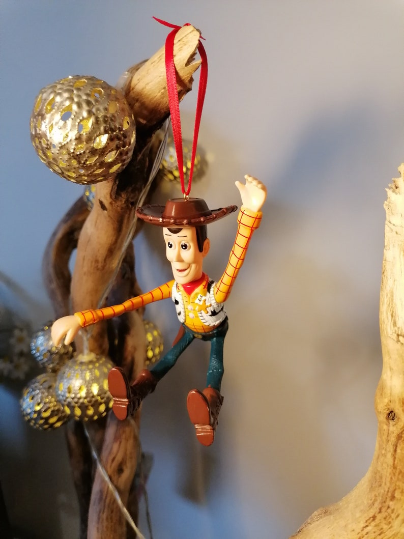 May include: A red-stringed ornament of Woody from Toy Story, wearing his signature cowboy hat and vest, with a yellow shirt and blue jeans. He is posed with his arms outstretched and legs bent, as if he is jumping or flying.