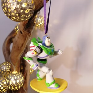K&ouml;nnte beinhalten: Ein gr&uuml;ner und wei&szlig;er Buzz Lightyear Weihnachtsbaumschmuck, der auf einem gelben Surfbrett steht, mit einem violetten Band befestigt.