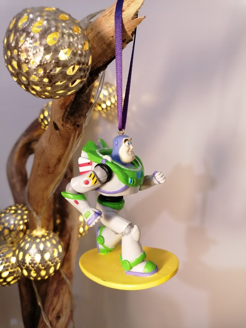 K&ouml;nnte beinhalten: Ein gr&uuml;ner und wei&szlig;er Buzz Lightyear Weihnachtsbaumschmuck, der auf einem gelben Surfbrett steht, mit einem violetten Band befestigt.