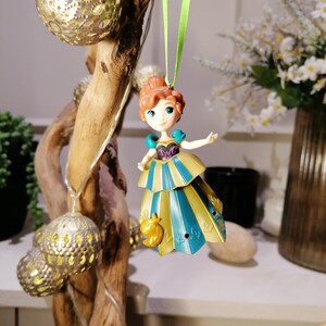 May include: A small plastic figurine of a princess wearing a blue and gold dress with a bird ornament hanging from a green ribbon.