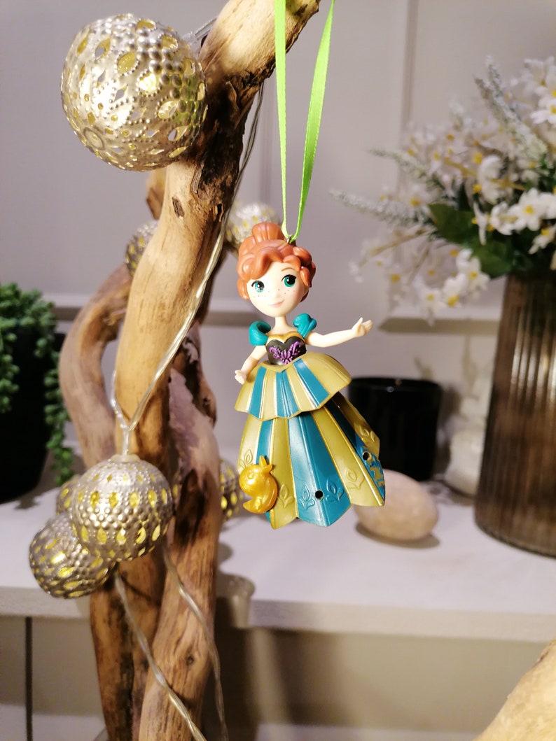 May include: A small plastic figurine of a princess wearing a blue and gold dress with a bird ornament hanging from a green ribbon.