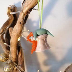May include: A green and purple Christmas bauble featuring a character from the Star Wars franchise, holding a bright orange object. The bauble is hanging from a green ribbon on a branch.