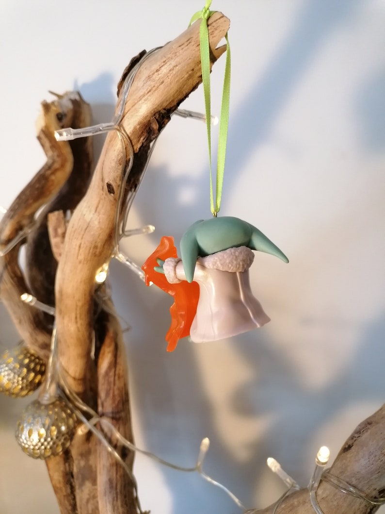 May include: A green and purple Christmas bauble featuring a character from the Star Wars franchise, holding a bright orange object. The bauble is hanging from a green ribbon on a branch.