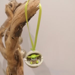 May include: A Christmas ornament featuring a small green character in a white pod, suspended by a light green ribbon. The character has large ears and is wearing a mask. The ornament is hanging from a piece of driftwood.