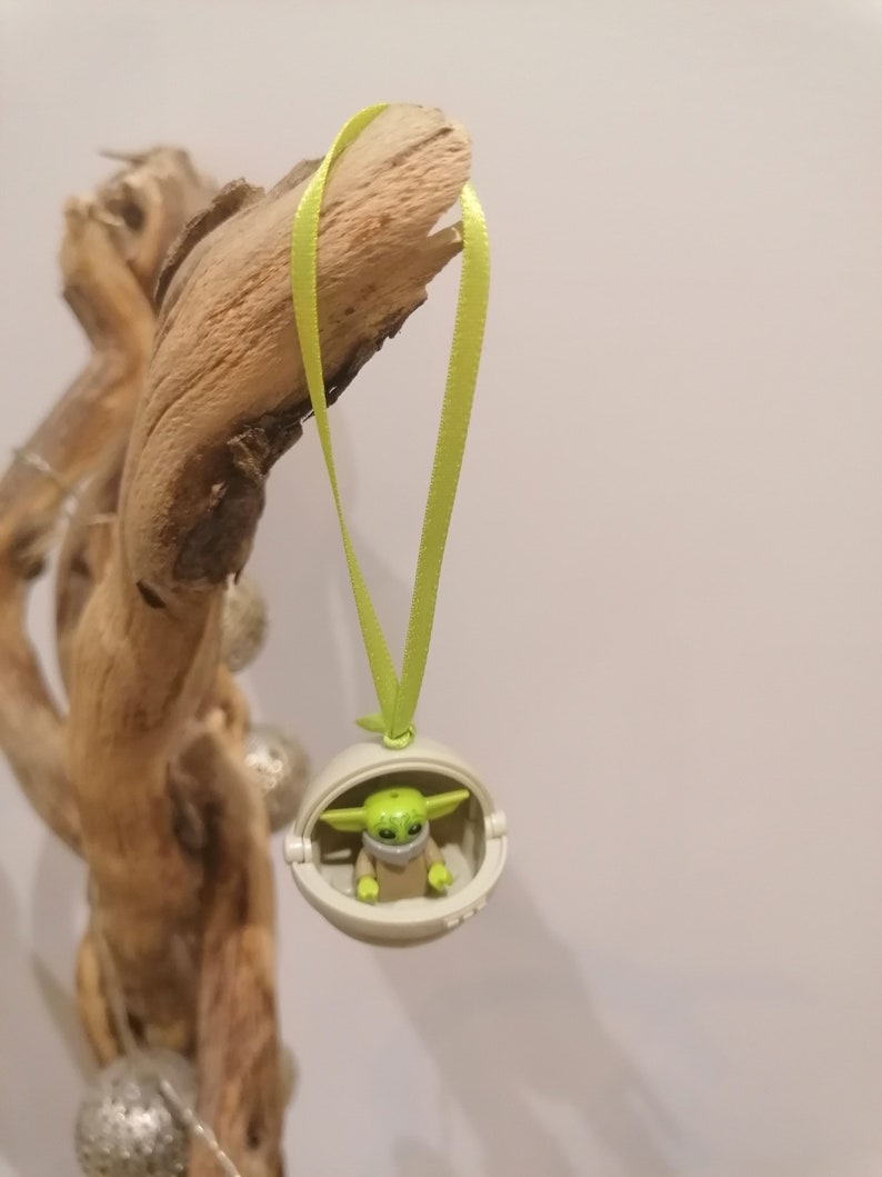 May include: A Christmas ornament featuring a small green character in a white pod, suspended by a light green ribbon. The character has large ears and is wearing a mask. The ornament is hanging from a piece of driftwood.