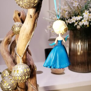 Peut inclure: Une d&eacute;coration en plastique bleue et blanche repr&eacute;sentant une princesse v&ecirc;tue d'une robe bleue avec un corsage blanc. La d&eacute;coration est suspendue &agrave; un ruban bleu sur une branche avec des guirlandes lumineuses dor&eacute;es.
