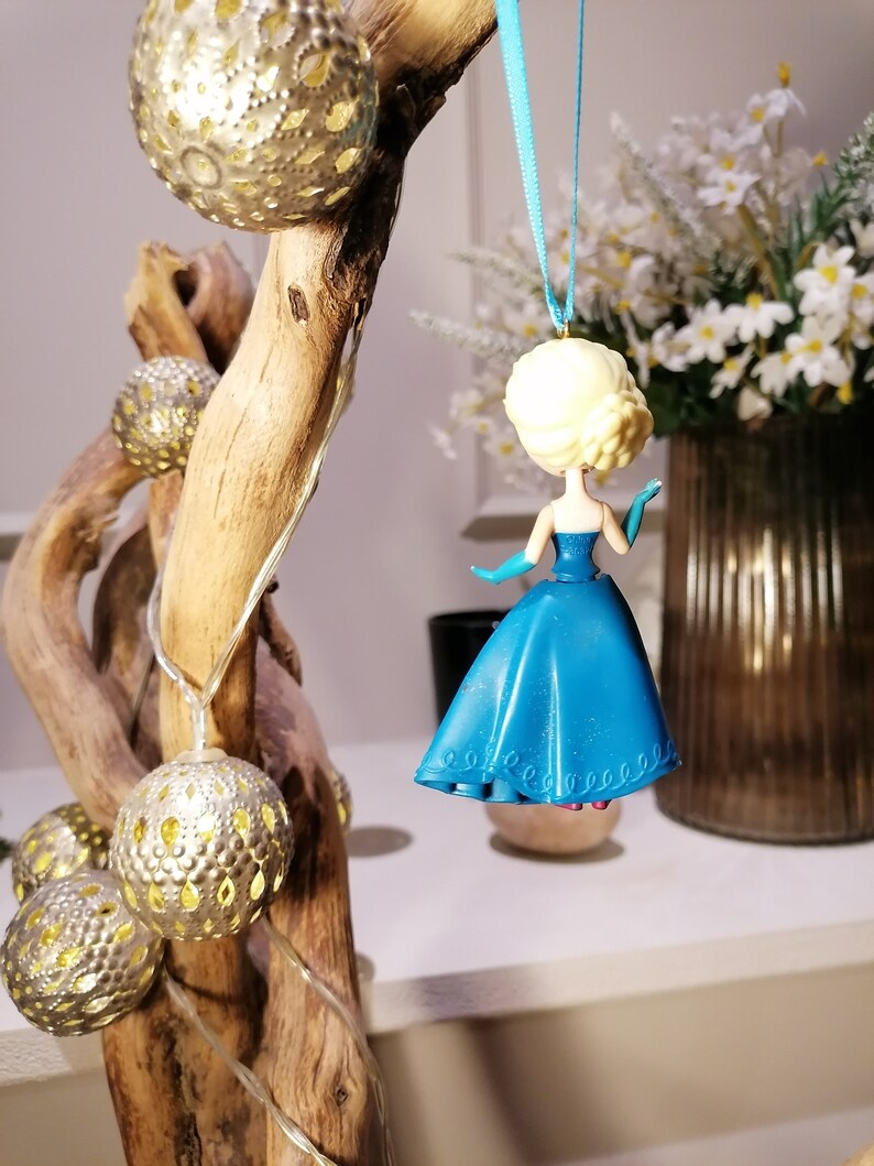 Peut inclure: Une d&eacute;coration en plastique bleue et blanche repr&eacute;sentant une princesse v&ecirc;tue d'une robe bleue avec un corsage blanc. La d&eacute;coration est suspendue &agrave; un ruban bleu sur une branche avec des guirlandes lumineuses dor&eacute;es.