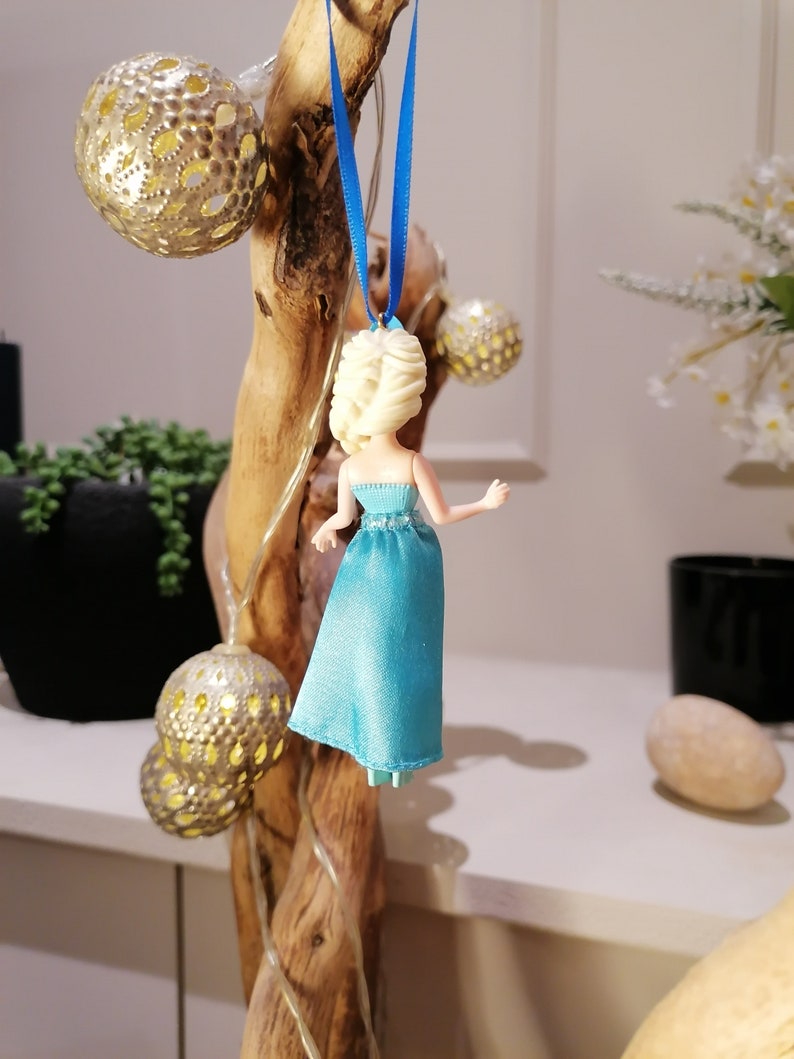 K&ouml;nnte beinhalten: Eine blaue und wei&szlig;e Plastikpuppe, die ein blaues Kleid mit wei&szlig;em Oberteil tr&auml;gt. Die Puppe h&auml;ngt an einem blauen Band an einem Holzast.