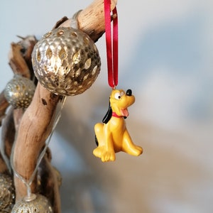 Op de afbeelding: Een gele en zwarte cartoonhond ornament hangend aan een rood lint op een tak met zilveren en gouden ornamenten.