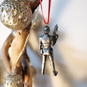 May include: A silver-coloured Christmas ornament featuring a figure holding a weapon. The ornament is hanging from a red ribbon.