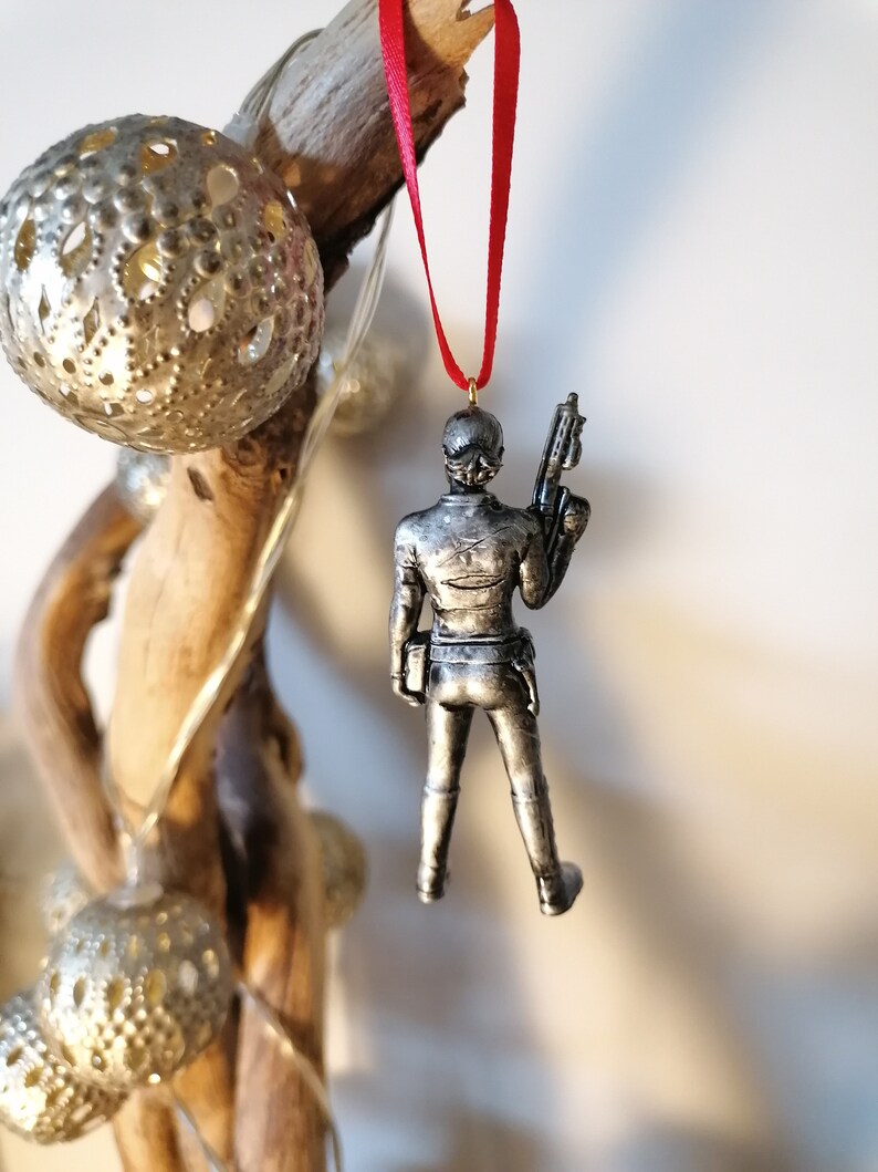May include: A silver-coloured Christmas ornament featuring a figure holding a weapon. The ornament is hanging from a red ribbon.
