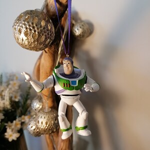K&ouml;nnte beinhalten: Eine Plastikfigur von Buzz Lightyear aus der Toy Story-Reihe. Die Figur tr&auml;gt seinen charakteristischen Weltraum-Ranger-Anzug in lila und gr&uuml;n. Die Figur h&auml;ngt an einem Ast mit einem lila Band.