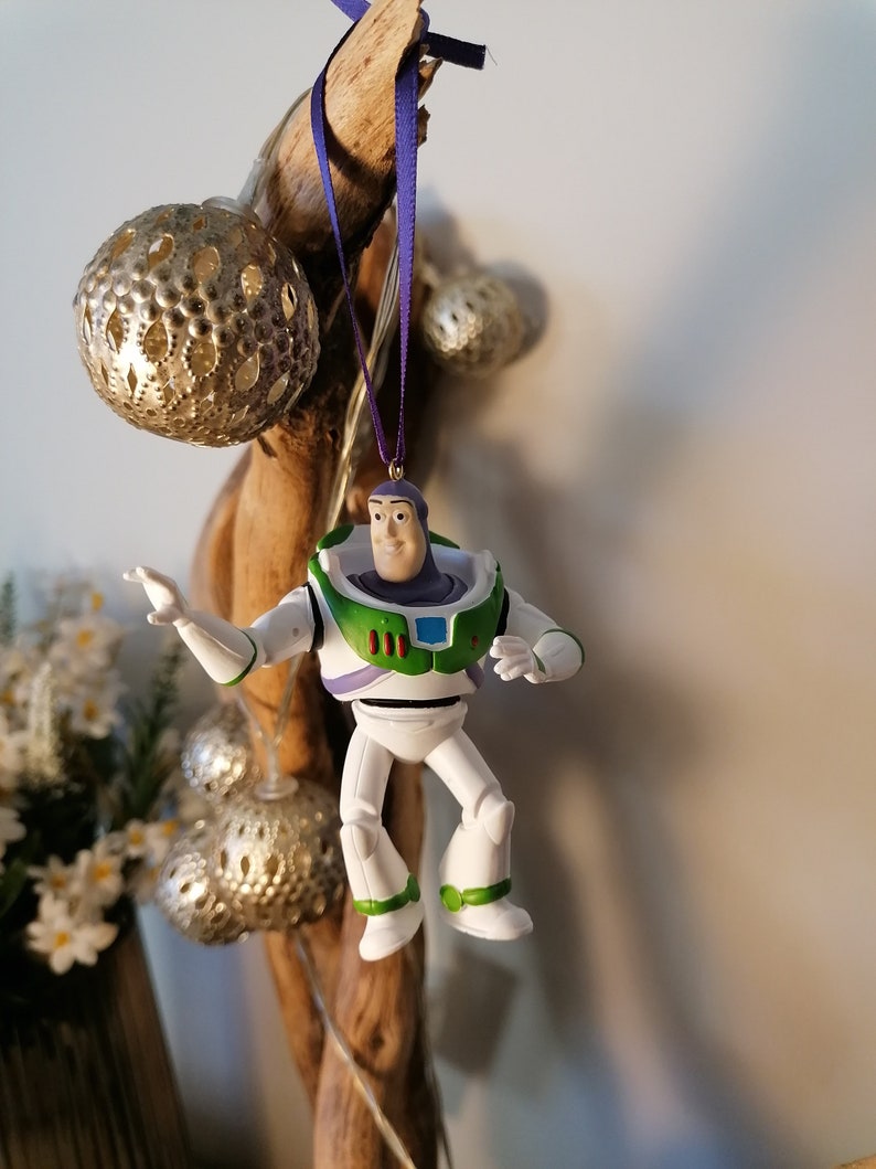 K&ouml;nnte beinhalten: Eine Plastikfigur von Buzz Lightyear aus der Toy Story-Reihe. Die Figur tr&auml;gt seinen charakteristischen Weltraum-Ranger-Anzug in lila und gr&uuml;n. Die Figur h&auml;ngt an einem Ast mit einem lila Band.