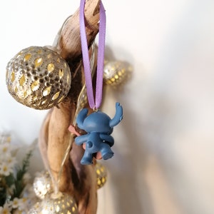 Op de afbeelding: Een blauwe Stitch-figuur ornament dat hangt aan een paars lint. Het ornament is bevestigd aan een stuk drijfhout met lichtsnoeren.