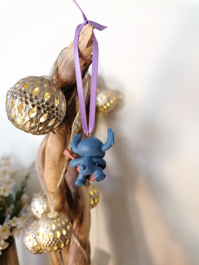 Op de afbeelding: Een blauwe Stitch-figuur ornament dat hangt aan een paars lint. Het ornament is bevestigd aan een stuk drijfhout met lichtsnoeren.