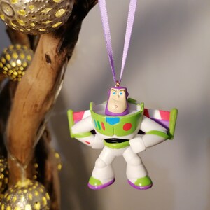 May include: A purple and green plastic toy ornament of Buzz Lightyear from the Toy Story franchise. The ornament is hanging from a purple ribbon.
