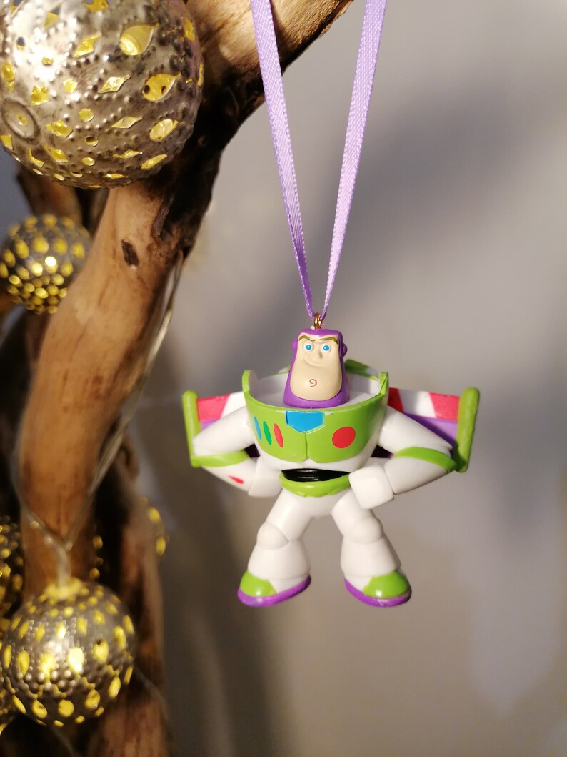 May include: A purple and green plastic toy ornament of Buzz Lightyear from the Toy Story franchise. The ornament is hanging from a purple ribbon.